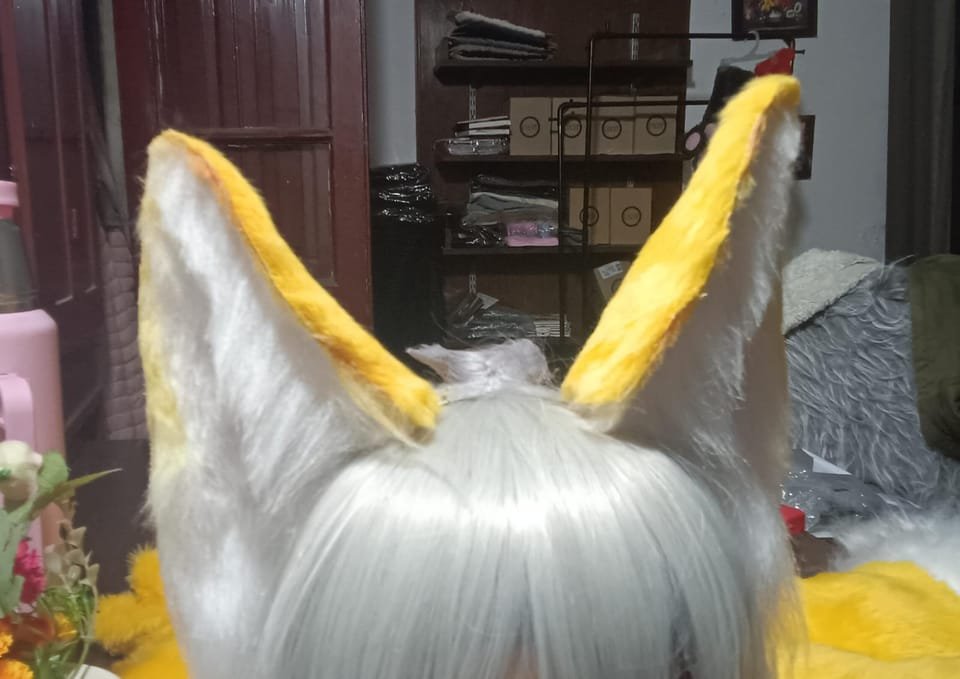 Vista trasera de una cabeza fursuit con orejas amarillas y blancas y peluca gris larga, en el taller.