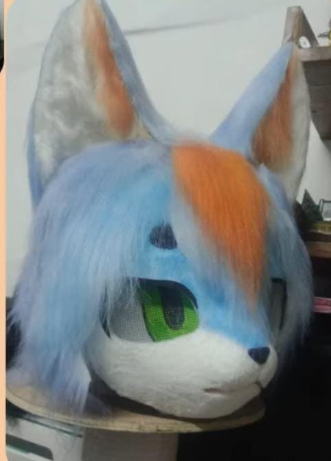Cabeza de fursuit zorro celeste y naranja con ojos verdes, vista de tres cuartos.