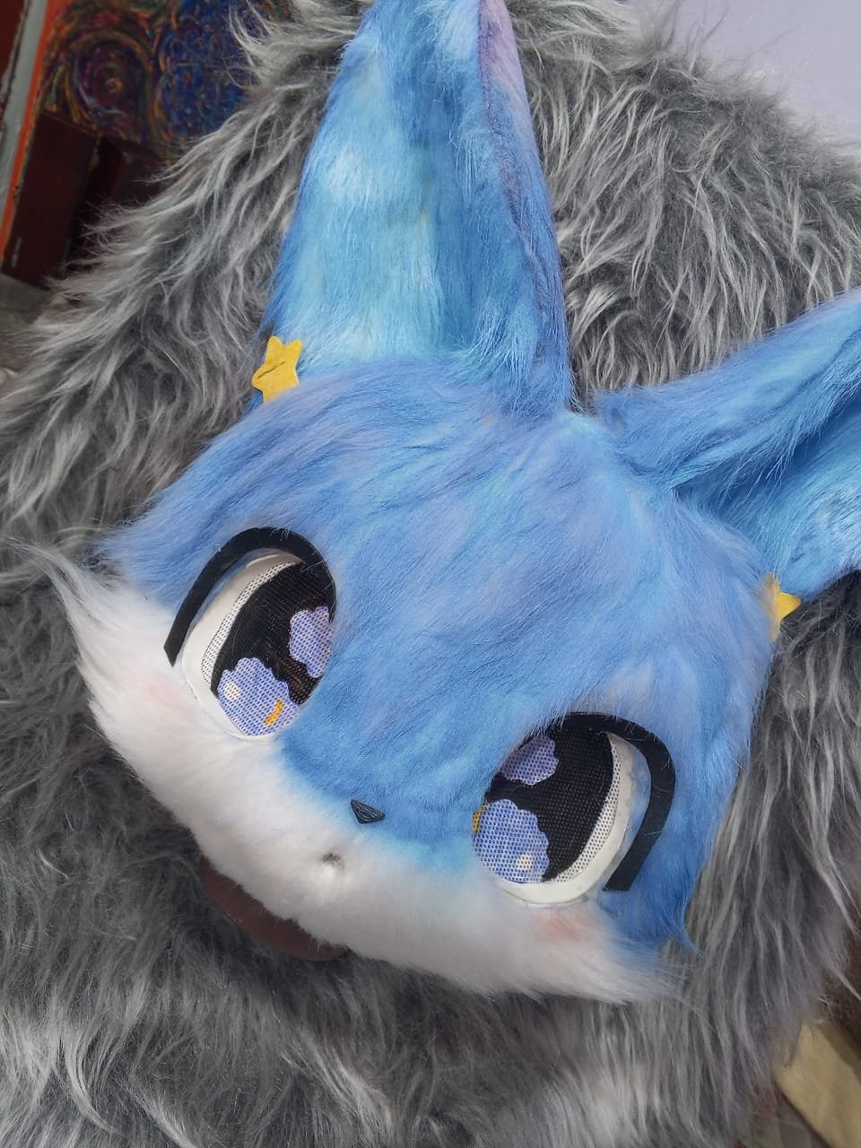 Cabeza de fursuit azul con orejas decoradas y ojos morados brillantes.